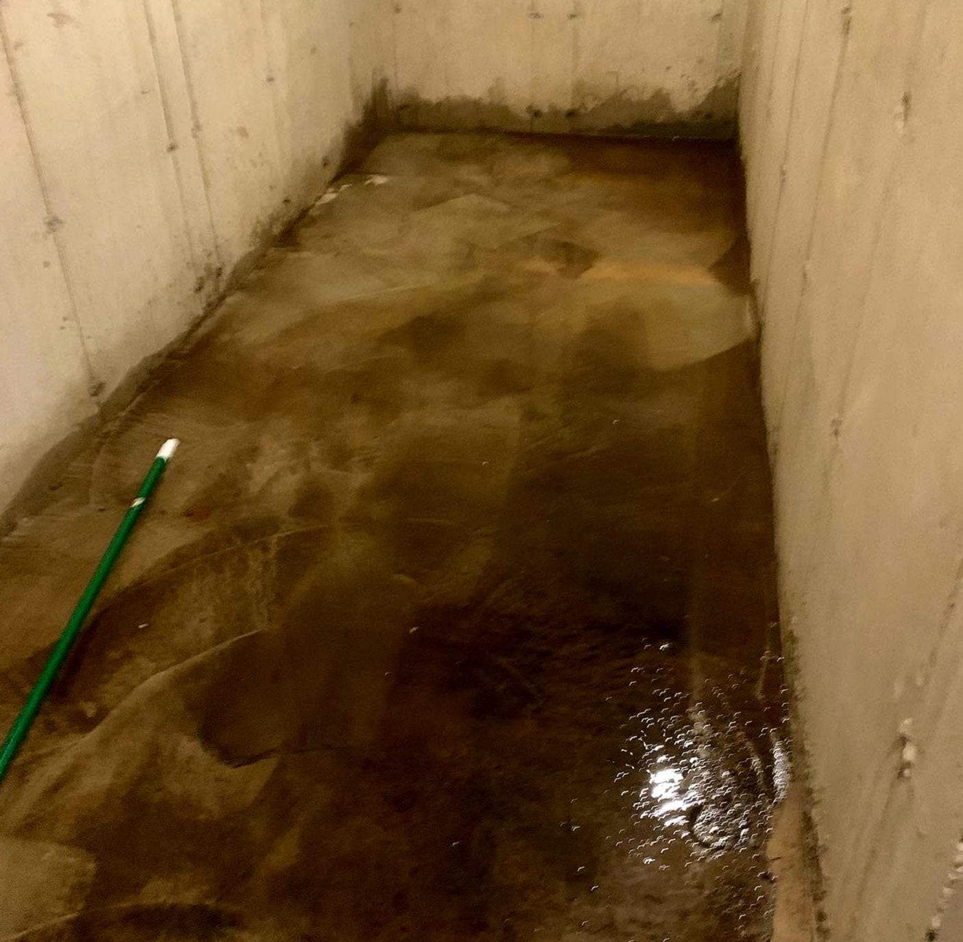 Muddy standing water across a concrete basement floor between poured concrete walls, dark staining along the bottom of the walls and a green broom handle lying in the water
