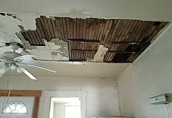A plaster ceiling broken away in large patches, exposing the wooden lath above it, with water stains on the remaining plaster and a white ceiling fan at the left