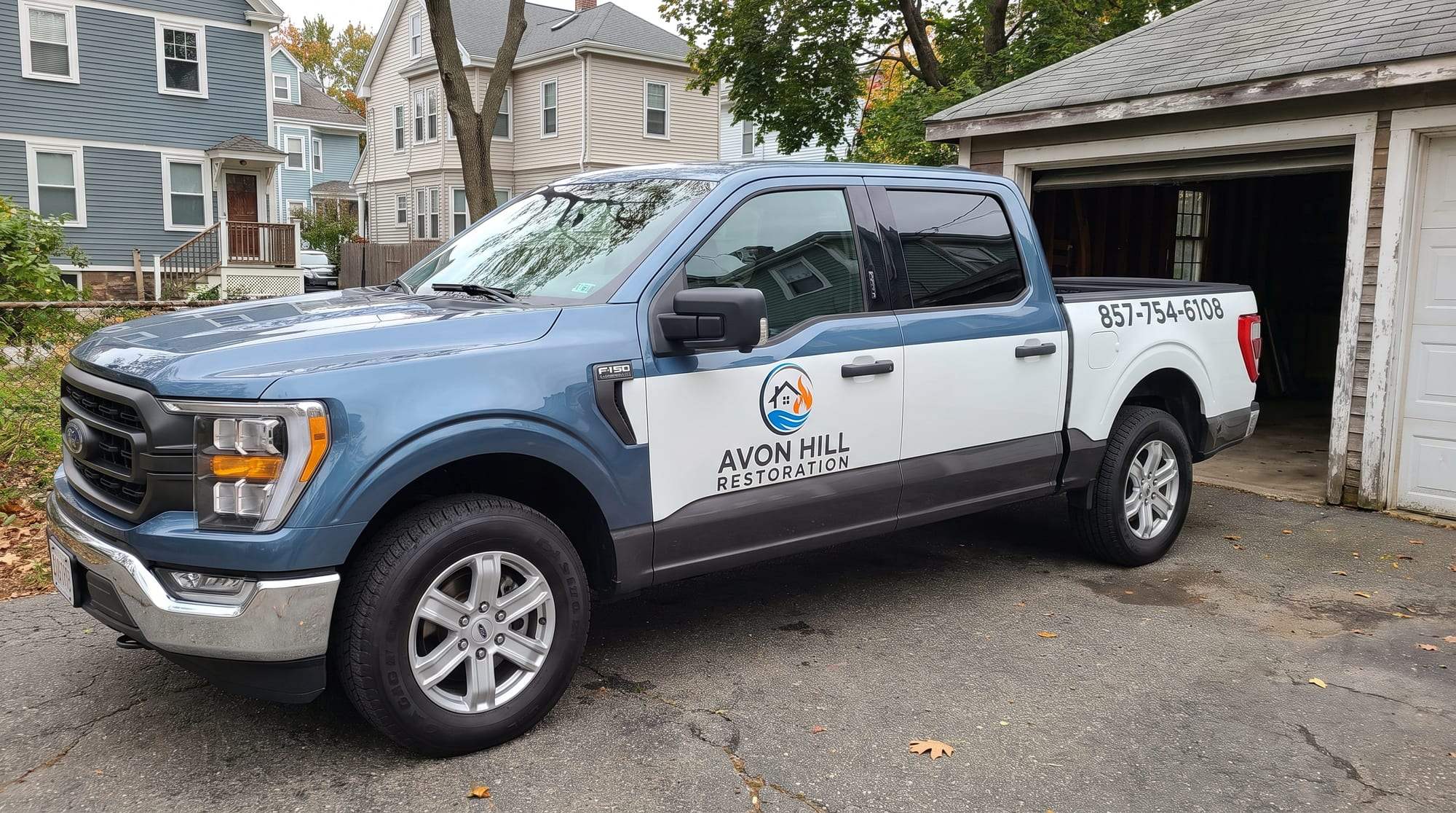 Avon Hill Restoration pickup truck, steel blue with white door and bed panels and a charcoal lower band, the company logo and name on the rear door and 857-754-6108 on the bed, parked on a driveway beside an open garage with clapboard houses and autumn trees behind