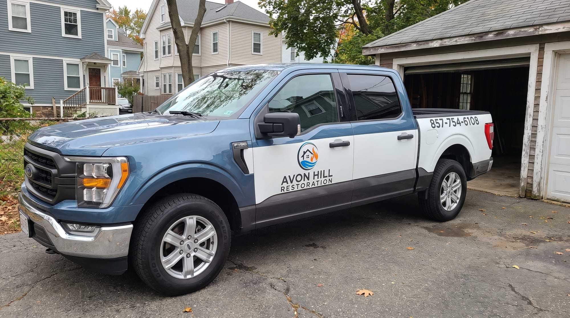 Avon Hill Restoration pickup truck, steel blue with white door and bed panels and a charcoal lower band, the company logo and name on the rear door and 857-754-6108 on the bed, parked on a driveway beside an open garage with clapboard houses and autumn trees behind