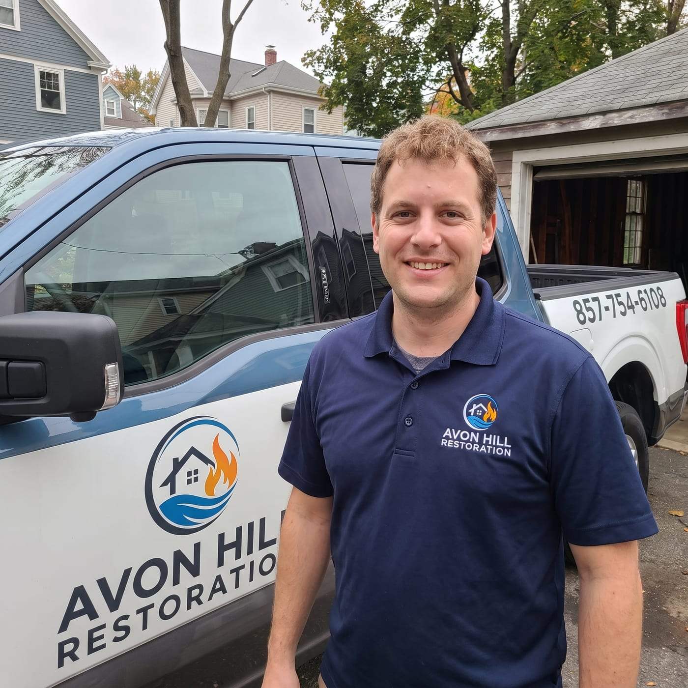 Spencer Burton, manager of Avon Hill Restoration, smiling in a navy polo shirt with the company logo, standing beside the company pickup truck in a residential driveway